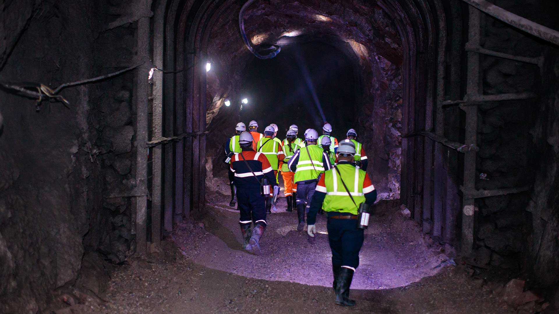 Group of worker in high visibility jackets entering a dark tunnel, low-lit with purple light.