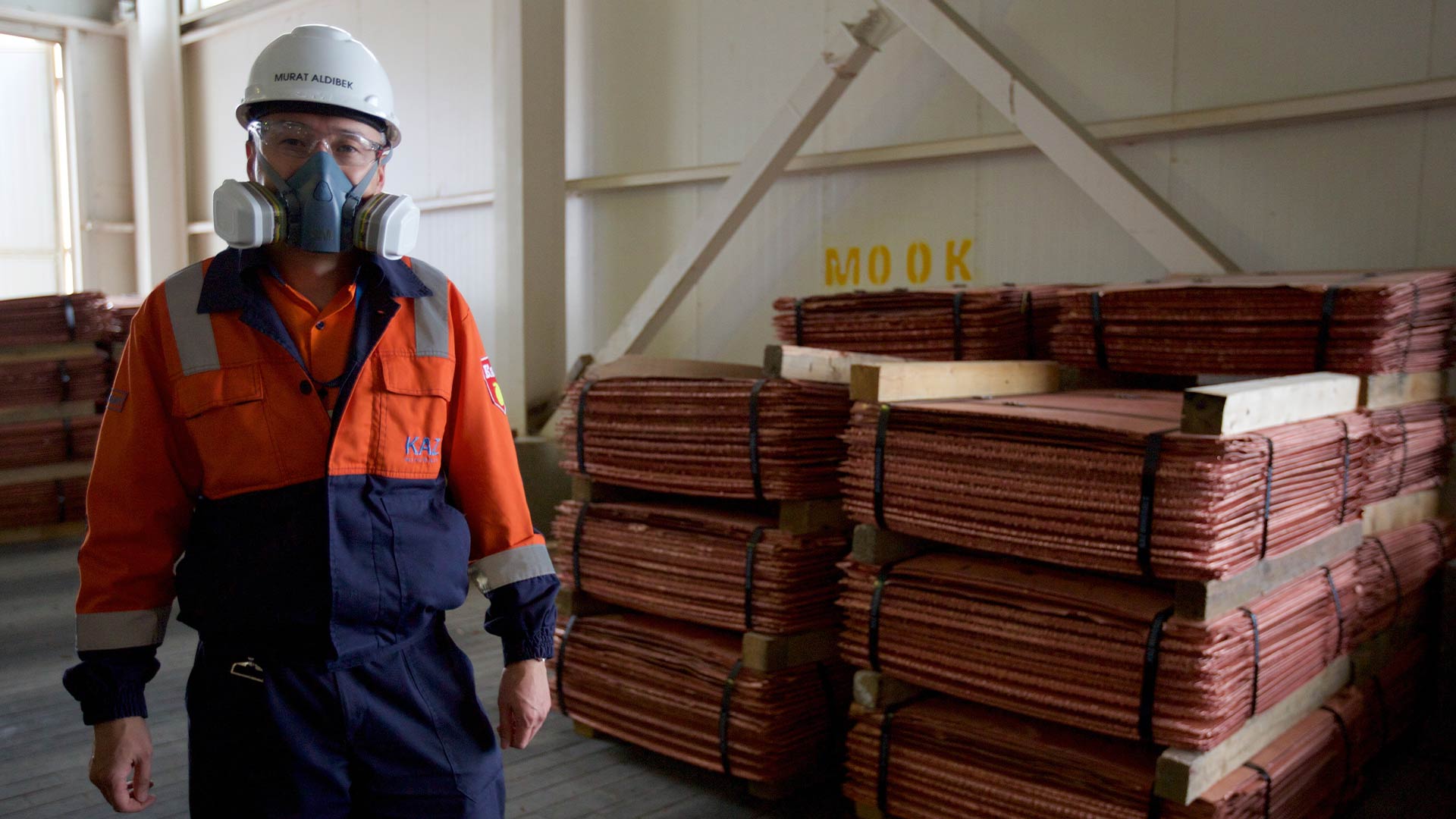 Қауіпсіздік құралын киген адам камераға қарап тұр. Материалдар фондық режимде жинақталған.