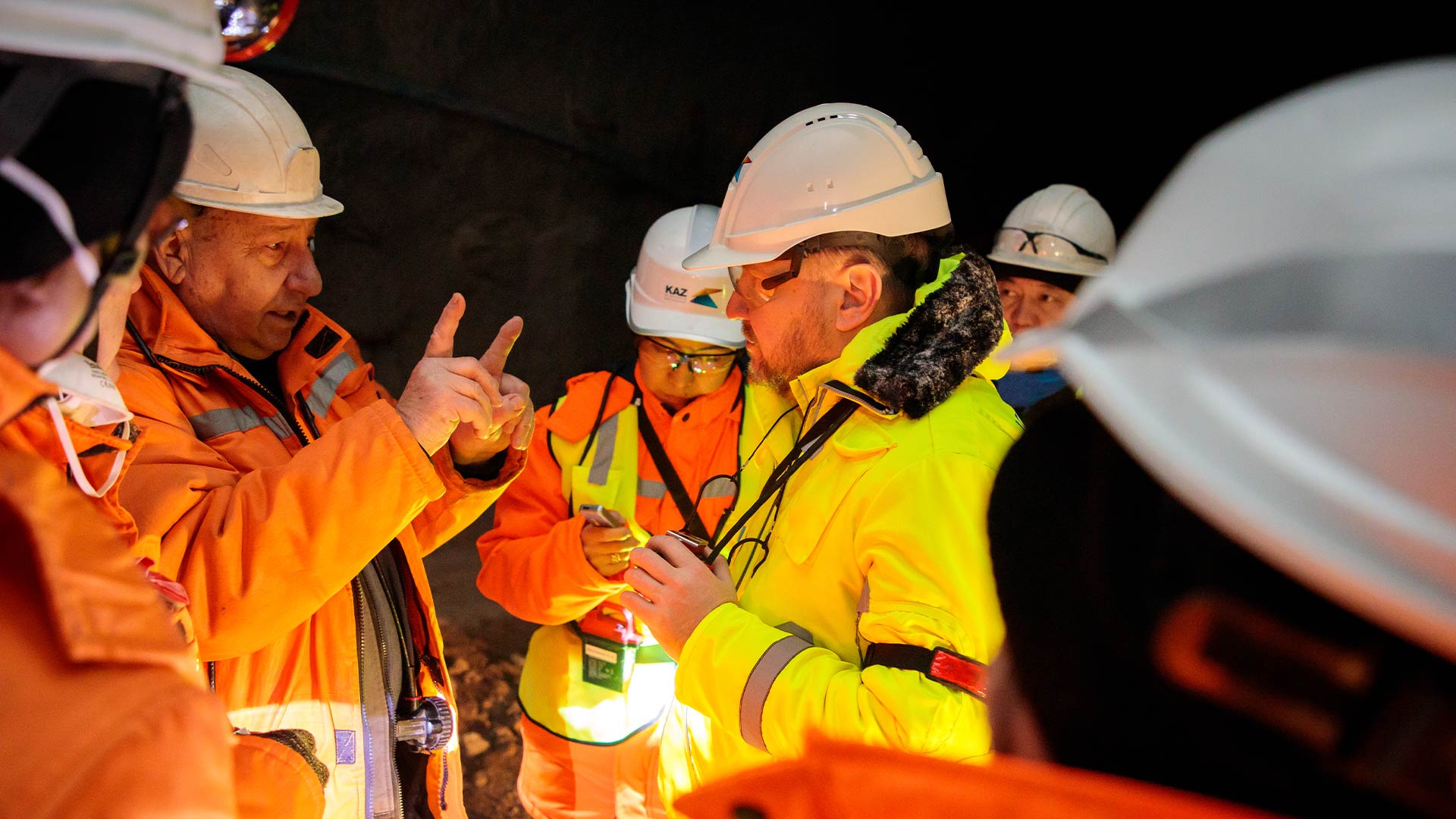 Үш жұмысшы алдыңғы қатарда қауіпсіздік киімдерін киіп, пікірталас жүргізеді. Артқы жағында қойма мен өңдеу орны бар.