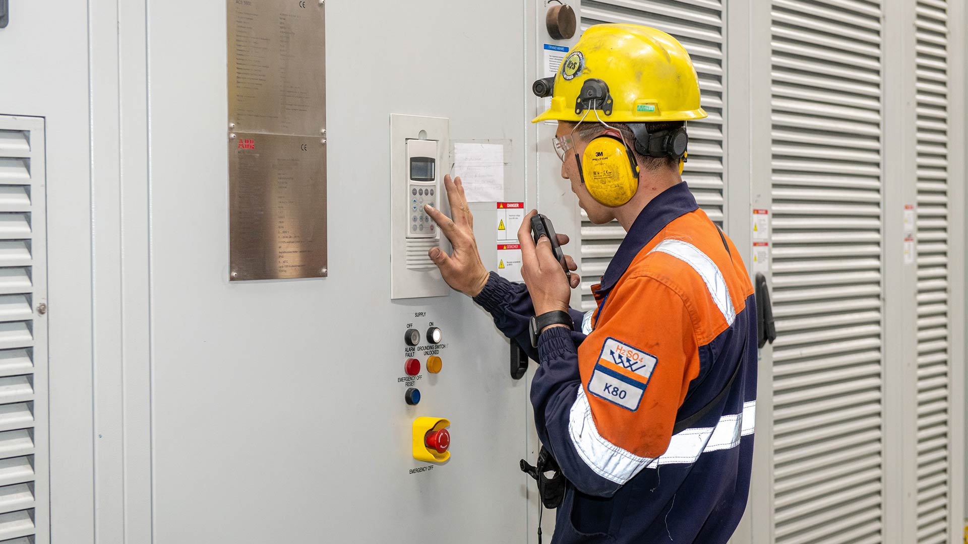 A worker in safety gear, including a yellow helmet and ear protection, communicates via a radio in front of a control panel, suggesting a focused and serious work environment.